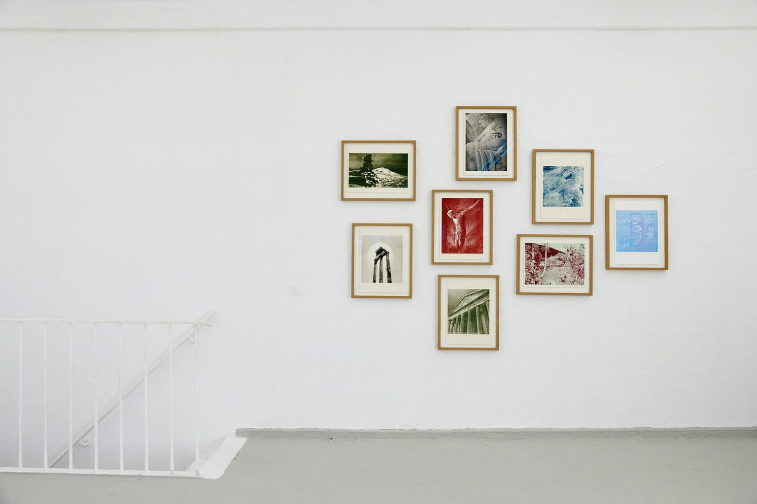Foto: Ein räum mit weißen Wänden, grauem Boden und Treppenaufgang. An der Wand hängt eine Gruppe aus gerahmten Objekten.