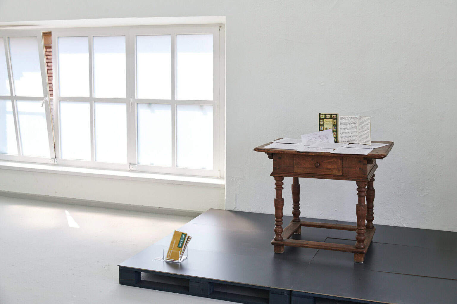 Ein Raum mit kleinem Fenster, weißen Wänden und grauem Boden. An der Wand steht ein schwarzer Podest, auf dem ein kleiner Tisch steht, auf dem Hefte und Zettel liegen.