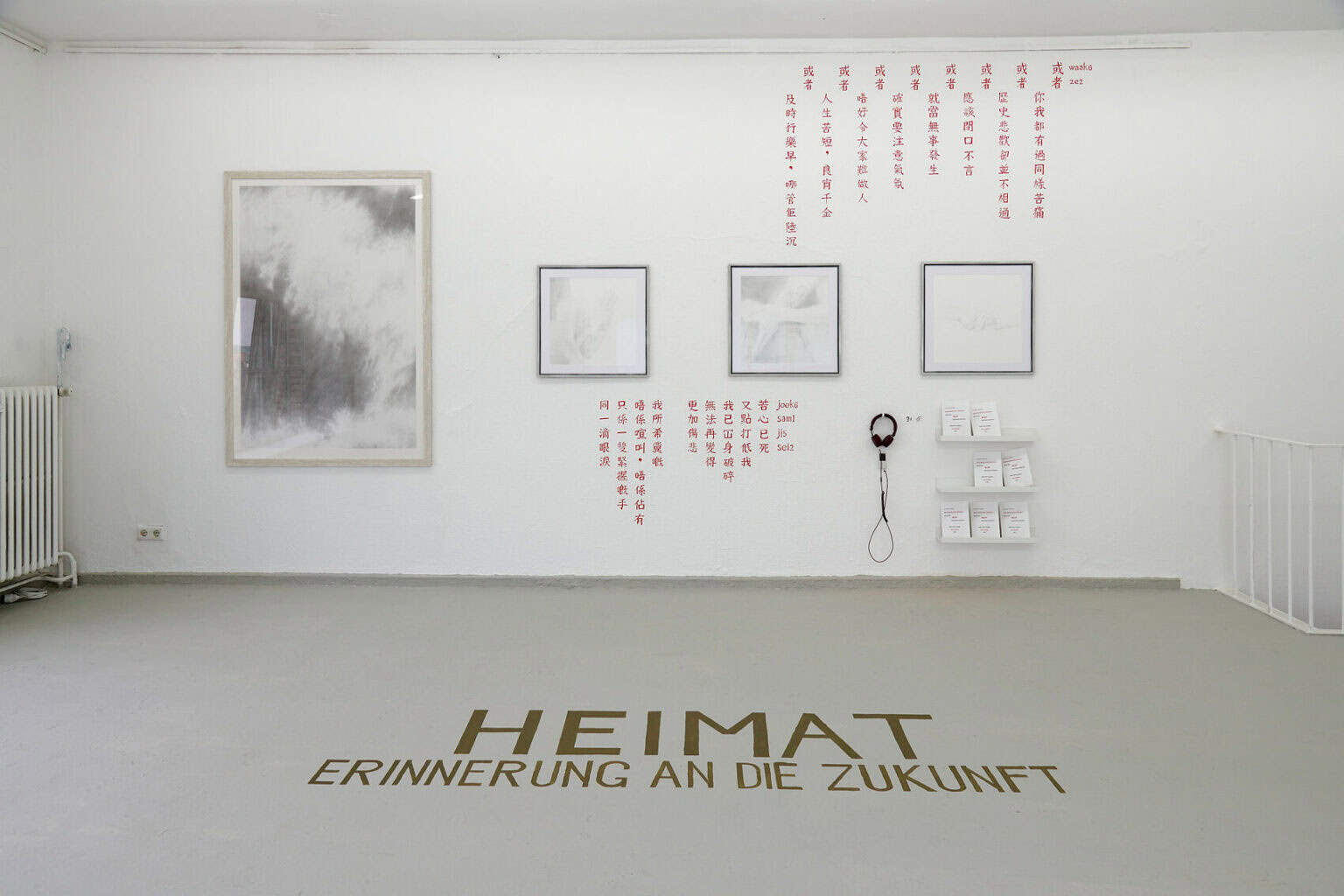 Foto: Ein Raum mit weißen Wänden und grauem Boden. Auf dem Boden stehen die Worte :"Heimat, Erinnerung an die Zukunft". An der Wand befinden sich vier gerahmte Objekte, die auf einer Mittellinie hängen. Die Objekte sind von roten, chinesischen und lateinischen Schriftzeichen umgeben. An der Wand befindet sich außerdem ein Regal mit Heften und Kopfhörer.