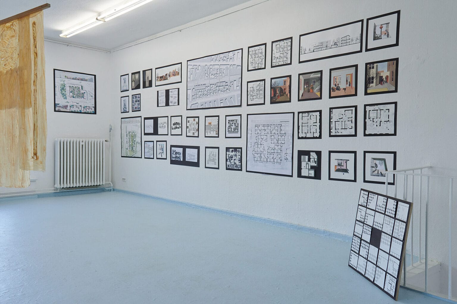 Foto: Ansicht eines Raumes mit weißen Wänden und hellblauem Boden. An den Wänden befinden sich eine Vielzahl von Papierarbeiten auf schwarzer Pappe. Links im Bild hängt ein an Seilen schwebendes Objekt von der Decke. Rechts im Bild steht ein flaches Objekt auf dem Boden, auf dem sich ebenfalls Papierarbeiten auf schwarzer Pappe befinden.