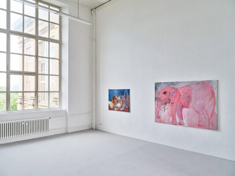 Foto: Aufnahme eines Ausstellungsraums mit weißen Wänden, grauem Boden und einem großen Fenster. An der rechten Wand hängen zwei abstrakte bunte Gemälde.