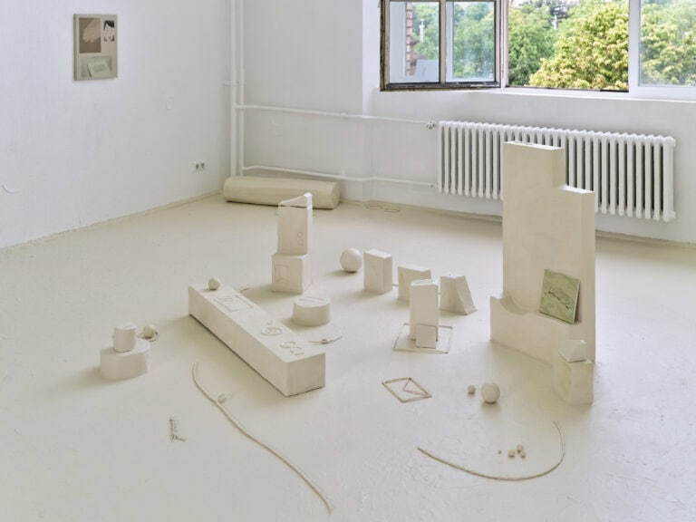 Foto: Aufnahme eines Ausstellungsraums mit weißen Wänden, beigem Boden und einem großen Fenster. Ein Fensterteil ist geöffnet. An der rechten Wand hängt ein abstraktes Gemälde. Auf dem Boden steht eine mehrteilige beige Installation.