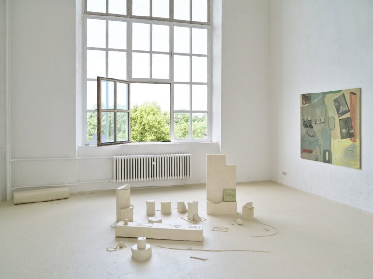 Foto: Aufnahme eines Ausstellungsraums mit weißen Wänden, beigem Boden und einem großen Fenster. Ein Fensterteil ist geöffnet. An der rechten Wand hängt ein abstraktes Gemälde. Auf dem Boden steht eine mehrteilige beige Installation.