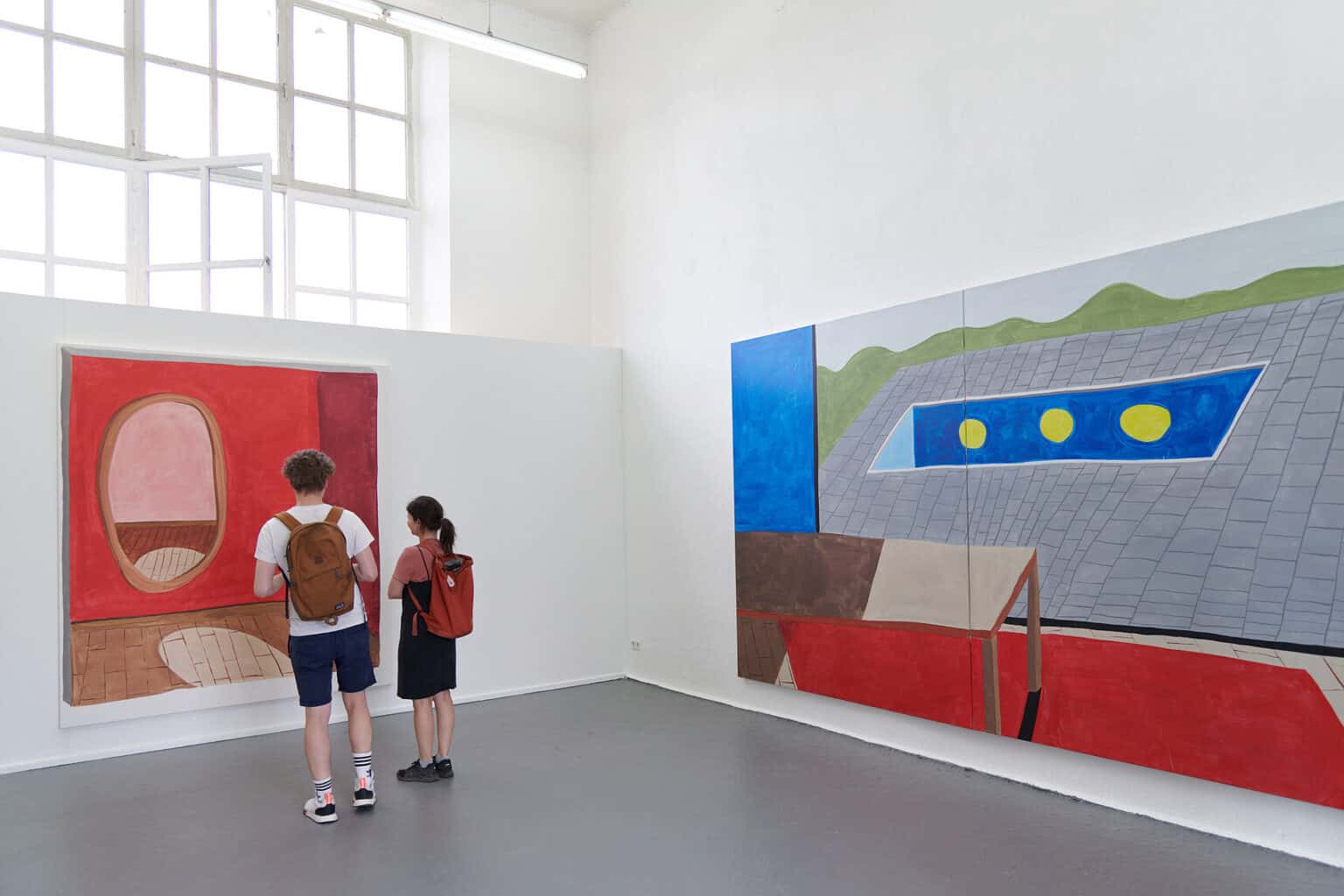 Foto: Ein Raum mit weißen Wänden und grauem Boden. Ein Teil des Fenster ist hinter einer eingezogenen Wand verborgen. An den Wänden hängen großformatige Leinwände. Eine der Arbeiten besteht aus zwei Leinwänden, die zusammen hängen und ein Bild ergeben. Im Raum stehen zwei Menschen.