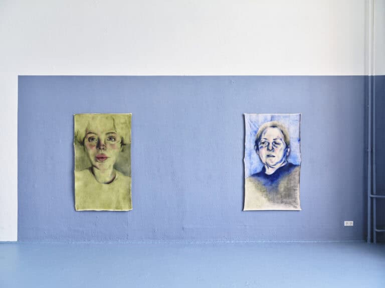 Foto: Aufnahme eines Ausstellungsraums mit weiß-blauen Wänden und blauem Boden. An der Wand hängen zwei Gemälde. Die Gemälde zeigen Porträts.