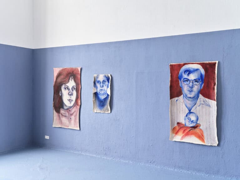 Foto: Aufnahme eines Ausstellungsraums mit weiß-blauen Wänden und blauem Boden. An der Wand hängen drei Gemälde. Die Gemälde zeigen Porträts.