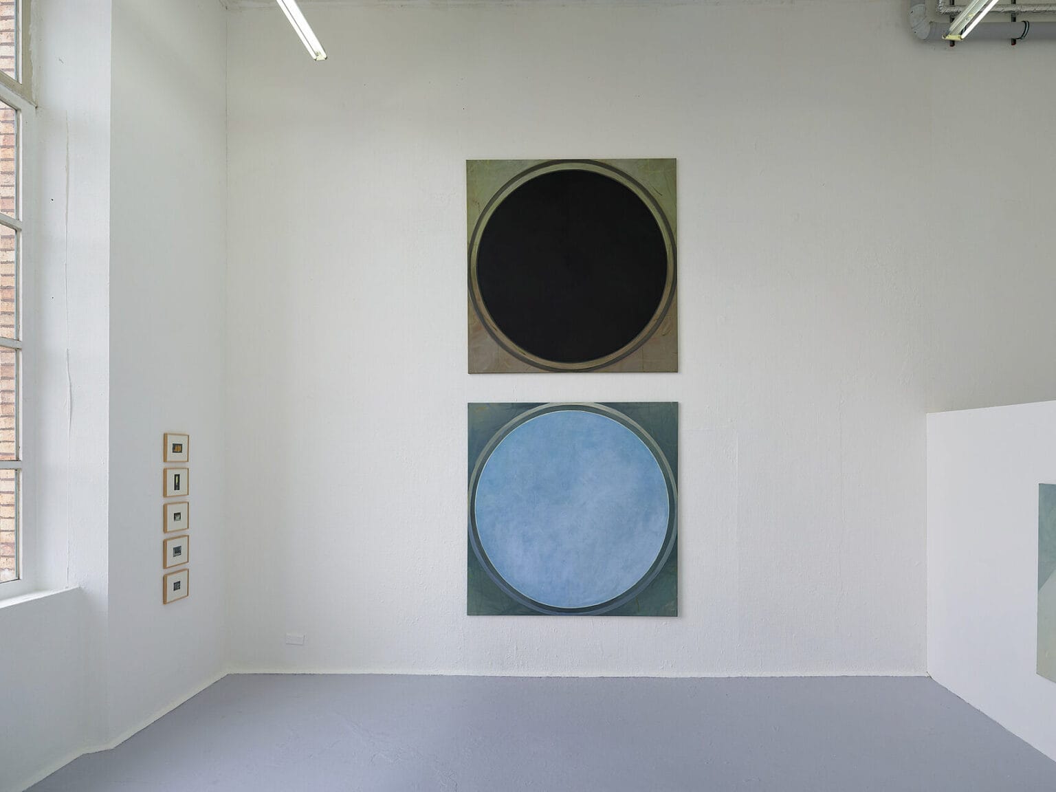 Foto: Ansicht eines Raumes mit weißen Wänden, grauem Boden und einem Fenster. Links an der Wand hängen in einer Linie untereinander mehrere gerahmte Arbeiten. An einer anderen Wand hängen zwei Leinwandarbeiten im gleichen Format untereinander.