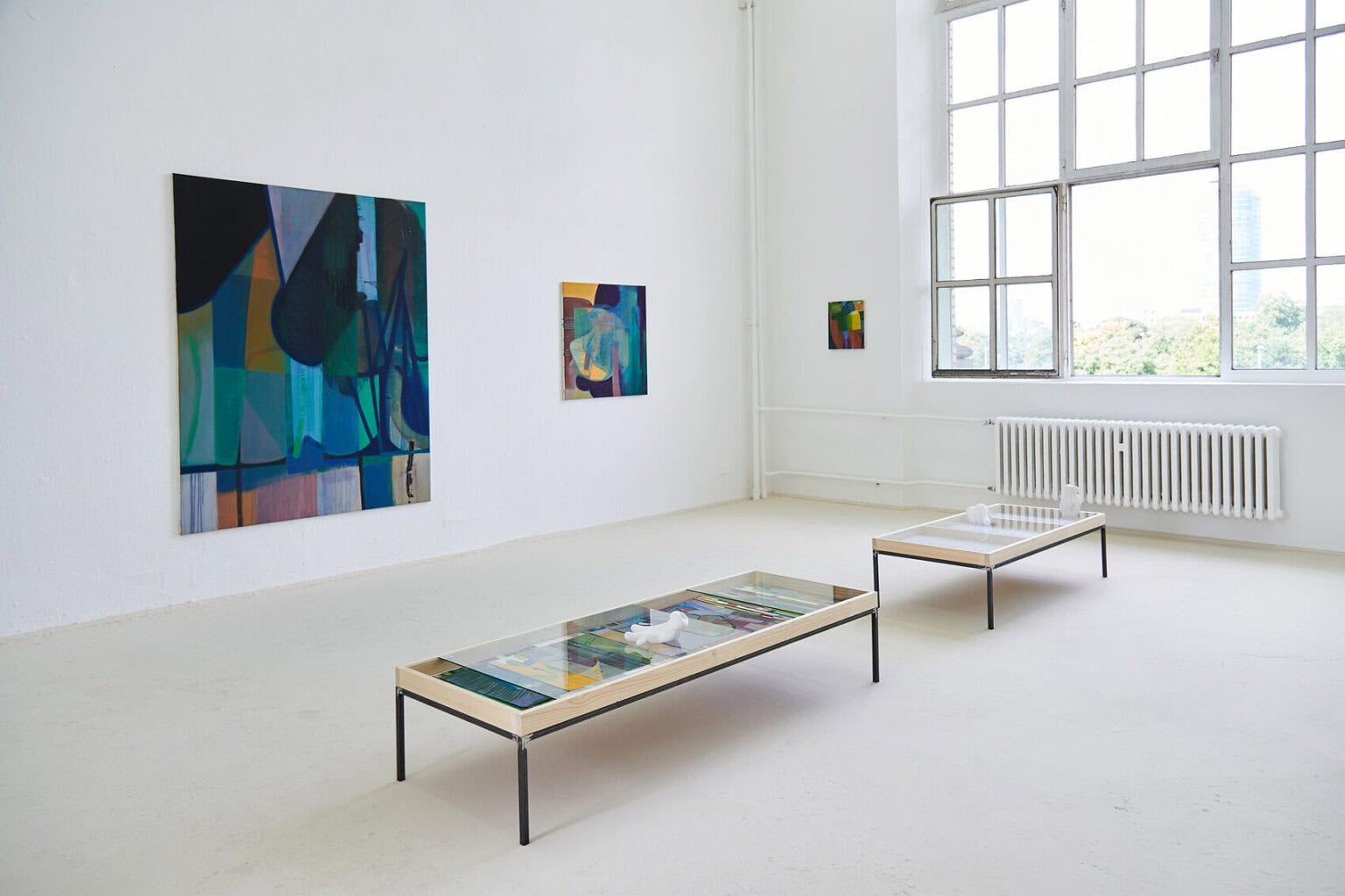 Foto: Ein Raum mit weißen Wänden und beigem Boden. Im Raum stehen zwei Vitrine, in denen sich Papierarbeiten befinden. Auf dem Glas der Vitrinen liegen Objekte. An den Wände hängen Leinwände im Klein-, Mittel- und Großformat.