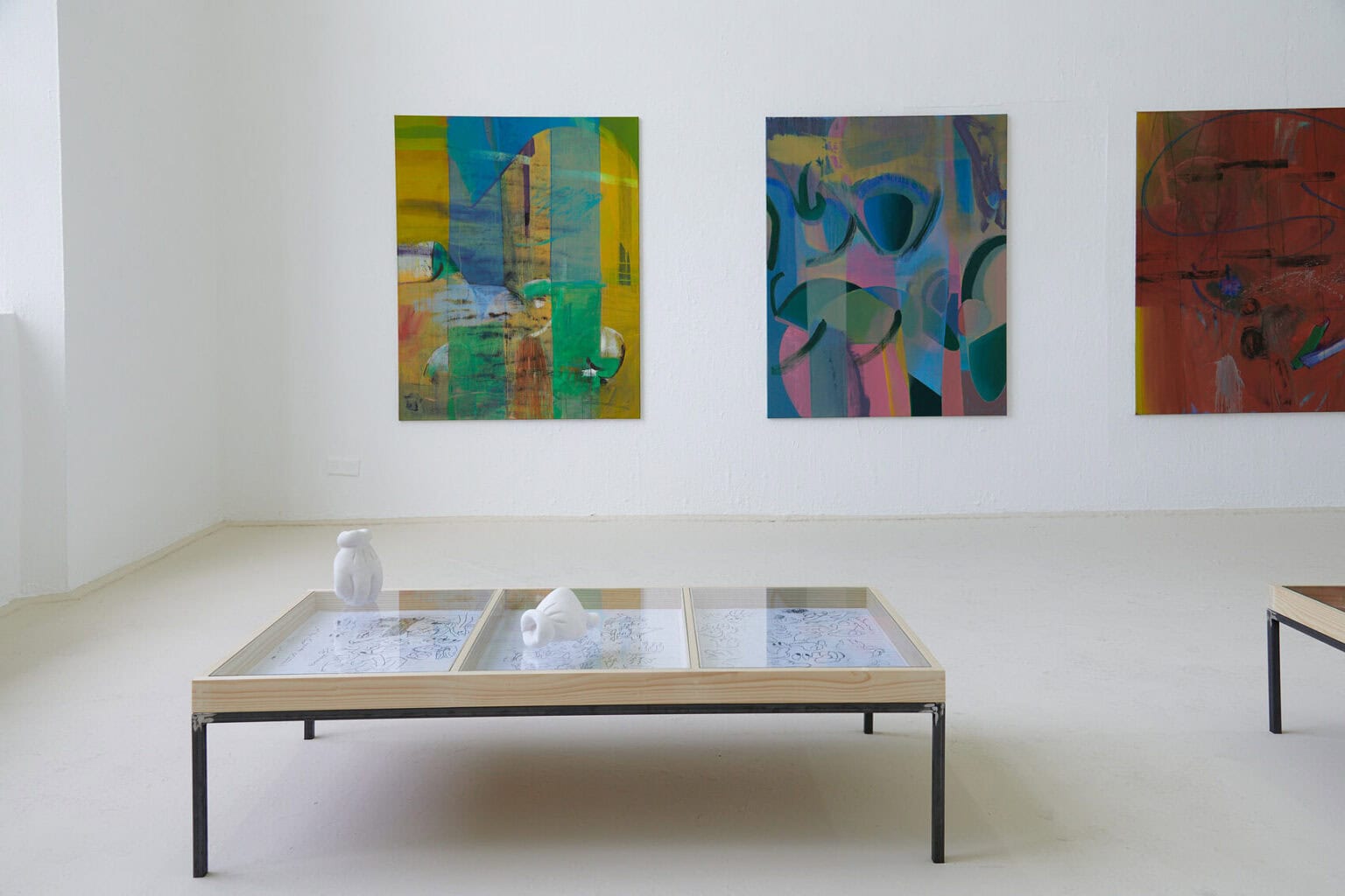 Foto: Ein Raum mit weißen Wänden und beigem Boden. Im Raum stehen zwei Vitrinen. In einer von ihnen liegen Papierarbeiten. Auf dem Glas der Vitrine, über den Zeichnungen, liegen zwei Objekte. An einer der Wände hängen drei großformatige Leinwandarbeiten.