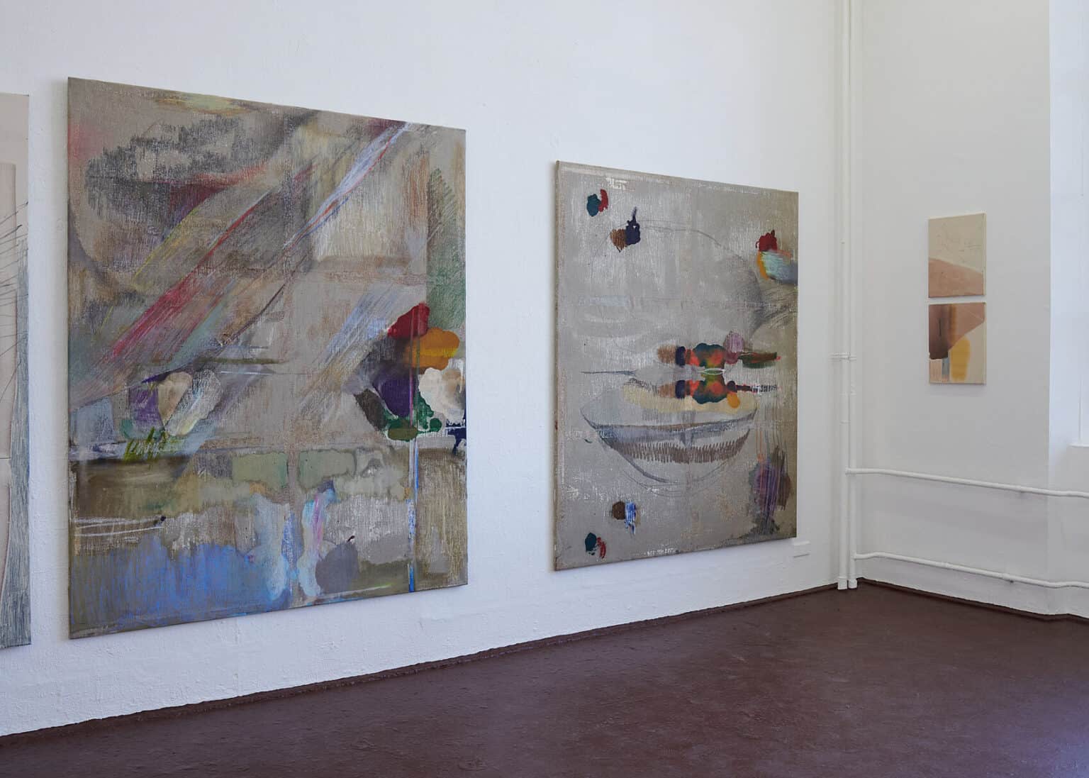 Foto: Ansicht eines Raumes mit weißen wänden und bräunlich-rotem Boden. An den Wänden hängen Malereien im Klein- und Großformat.