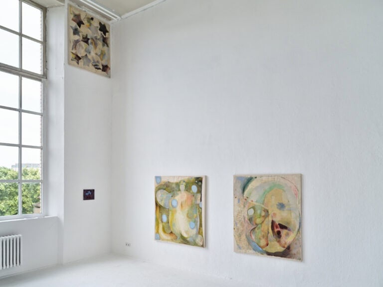Foto: Aufnahme eines Ausstellungsraums mit weißen Wänden, weißem Boden und einem großen Fenster. An den Wänden hängen vier abstrakte bunte Gemälde.