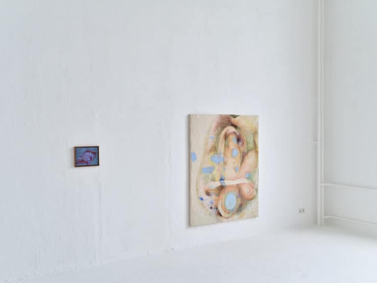 Foto: Aufnahme eines Ausstellungsraums mit weißen Wänden und weißem Boden. An den Wänden hängen zwei abstrakte bunte Gemälde in unterschiedlichen Größen.