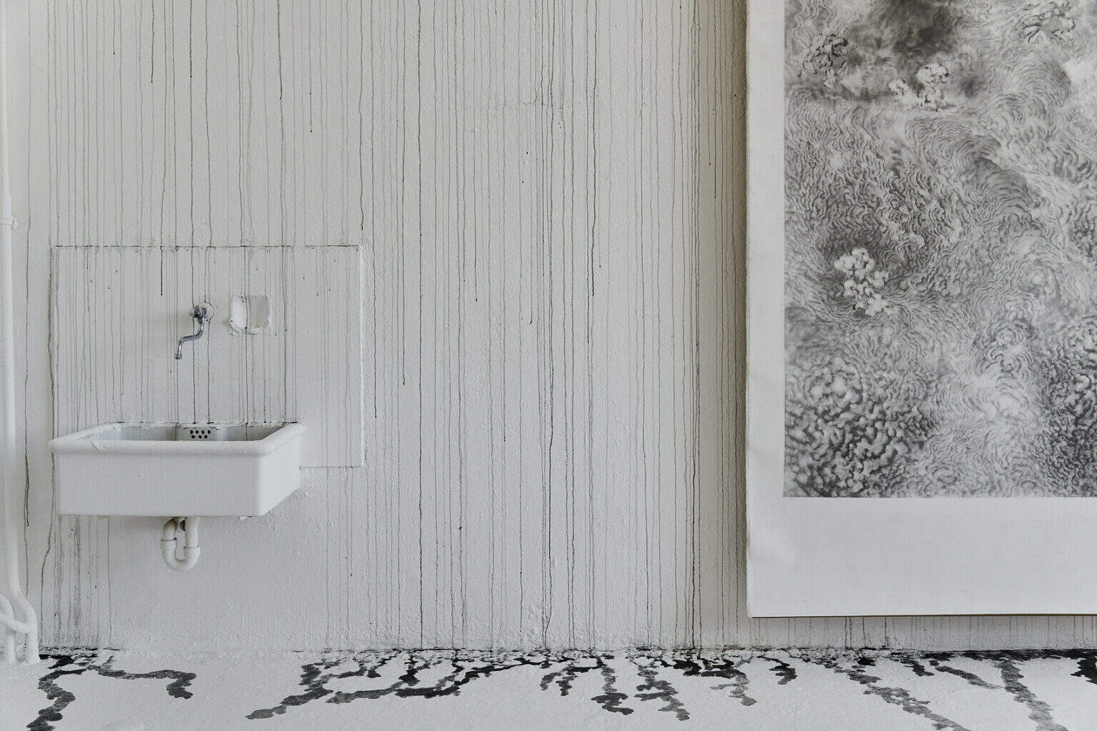 Foto: Ein Raum mit weißen Wänden und grauem Boden. Über die gesamte Länge der weißen Wand ziehen sich Laufspuren schwarzer Farbe, die beim Auftreffen auf dem Boden kleine Lachen bilden. Der Streifen des Bodens, auf dem sich die Lachen abzeichnen, ist im unterschied zum Rest des Bodens in einem Wandnahen Streifen ebenfalls weiß gestrichen. An der Wand hängt eine großformatige, monochrome Arbeit auf Papier. An der Wand befindet sich außerdem ein Waschbecken.