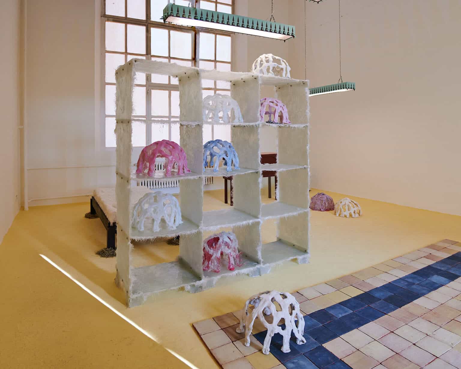 Foto: Ansicht eines Raumes mit weißen Wänden, gelbem Boden und einem großen Fenster. Von der Decke hängen zwei längliche Deckenlampen mit gemusterter Rahmung. Im Raum stehen mehrere Objekte aus unterschiedlichen Materialien. Auf dem Boden liegen verschiedenfarbige Kacheln, die dicht aneinander liegen und in einer quadratischen Form angeordnet sind. Auf den Kacheln steht ebenfalls ein Objekt.