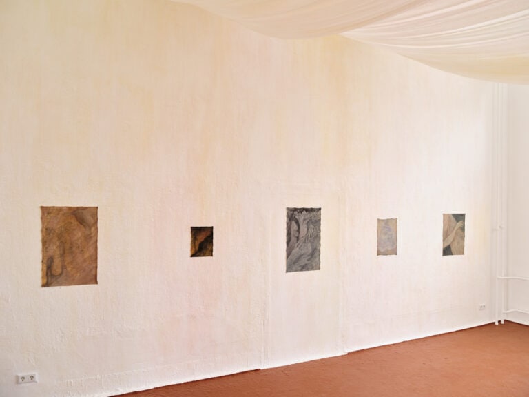 Foto: Aufnahme eines Ausstellungsraums mit hellen Wänden und braunem Boden. An der Decke ist mit Tüchern verhängt. An der Wand hängen fünf abstrakte Gemälde.