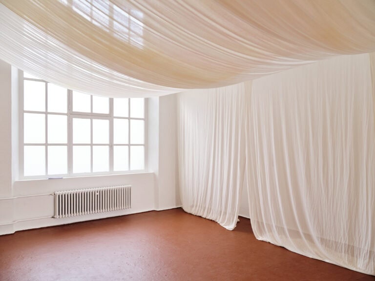 Foto: Aufnahme eines Ausstellungsraums mit weißen Wänden, braunem Boden und einem großen Fenster. Der Raum ist mit weißen Tüchern baldachinartig verhängt.