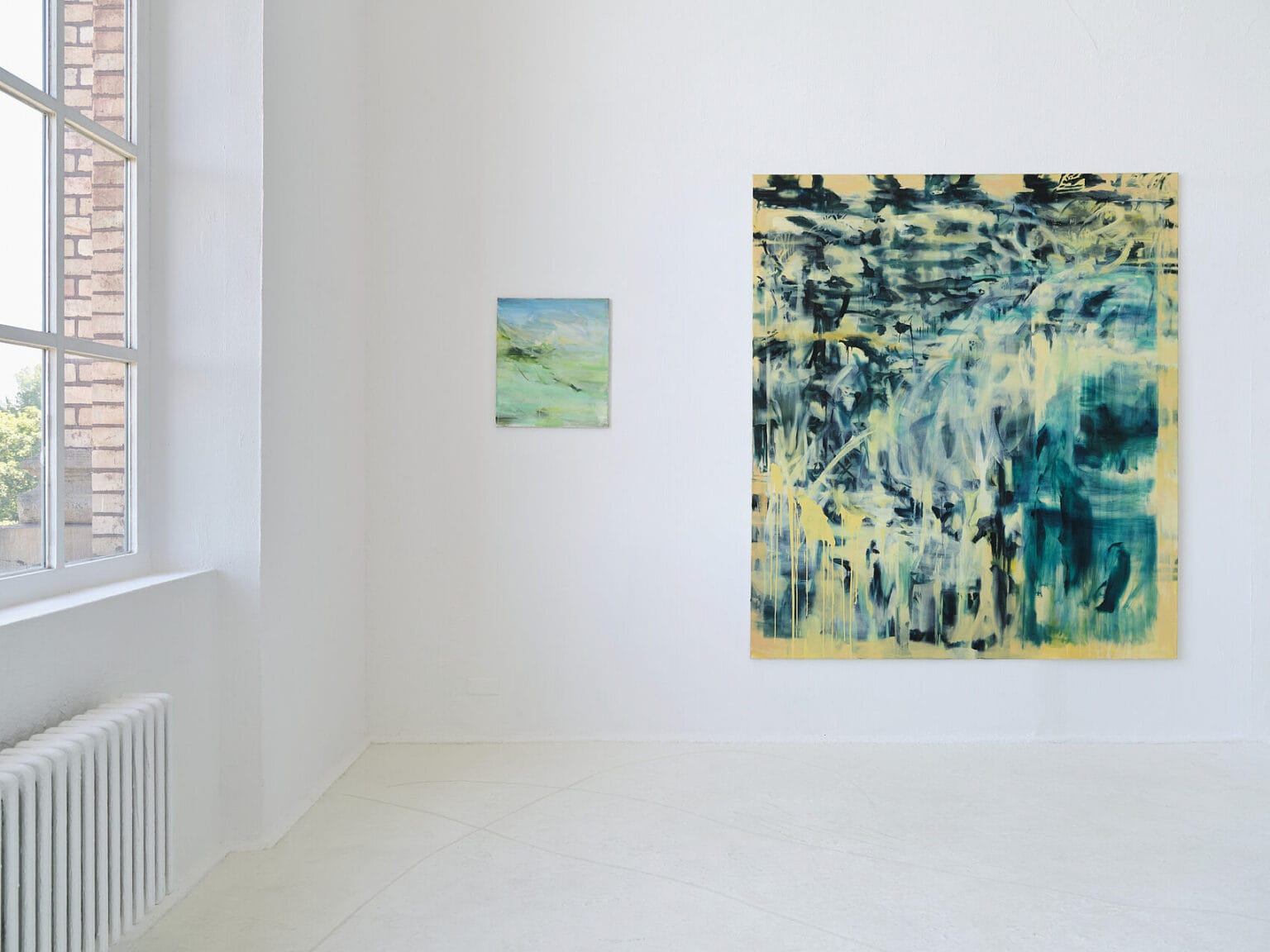 Foto: Ansicht eines Raumes mit weißen Wänden, hellgrauem Boden und einem Fenster. An der Wand hängen, auf unterschiedlichen Höhen, Leinwandarbeiten im Klein- und Großformat.