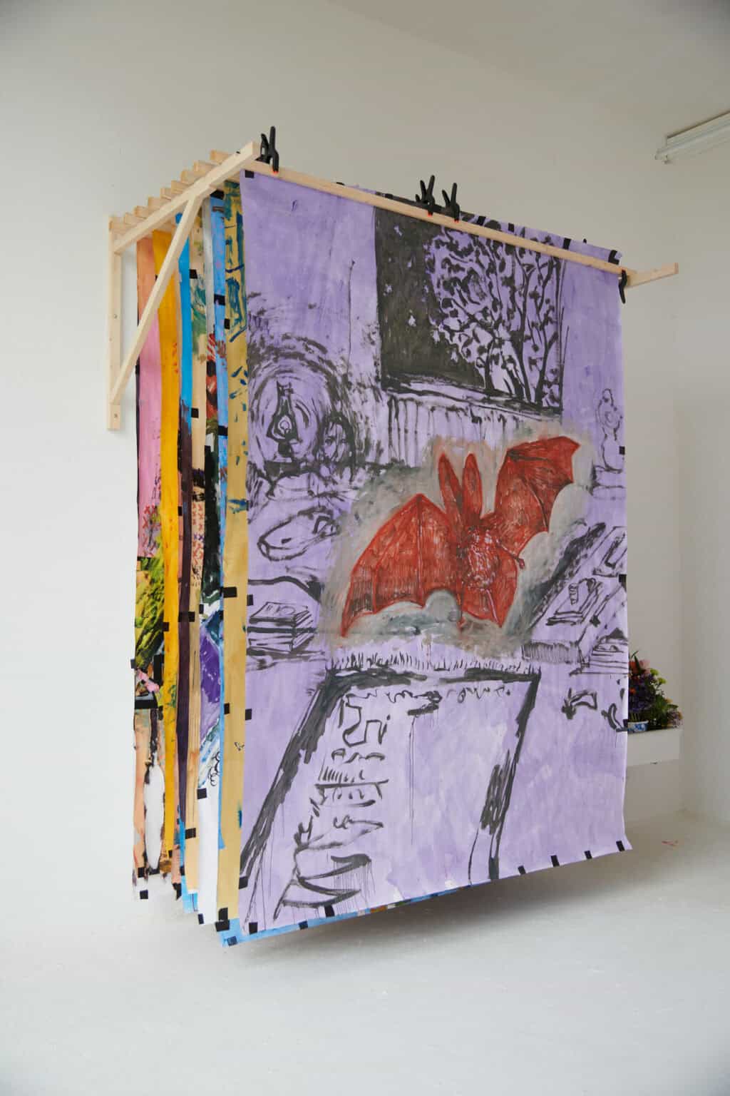 Foto: Ein Raum mit weißen Wänden und weißem Boden. Oben an der Wand befindet sich eine Holzkonstruktion, an der große Papierarbeiten hintereinander aufgehangen sind. Bloß die vorderste Arbeit ist gut zu sehen.