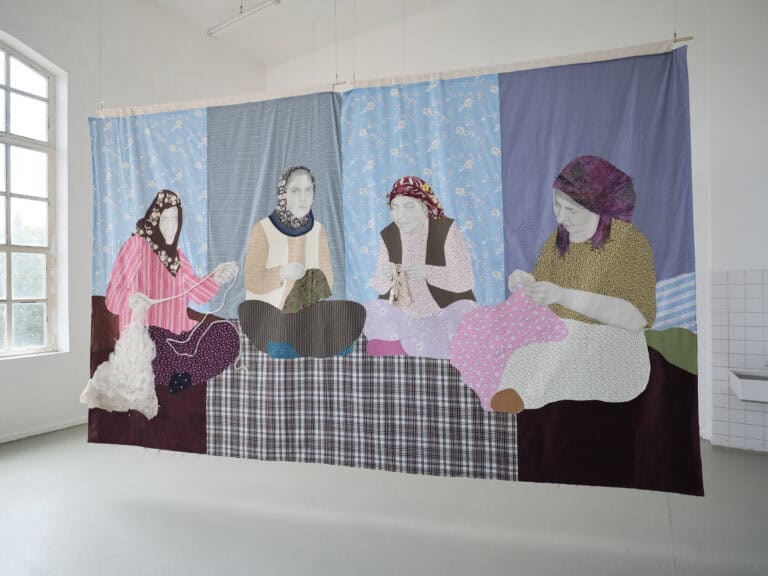 Foto: Aufnahme eines Ausstellungsraums mit weißen Wänden, grauem Boden und einem großen Fenster. Ein großformatiges Textilbild hängt von der Decke abgehängt. Das Bild zeigt vier weibliche Personen, die auf dem Boden Sitzen und Handarbeiten machen.
