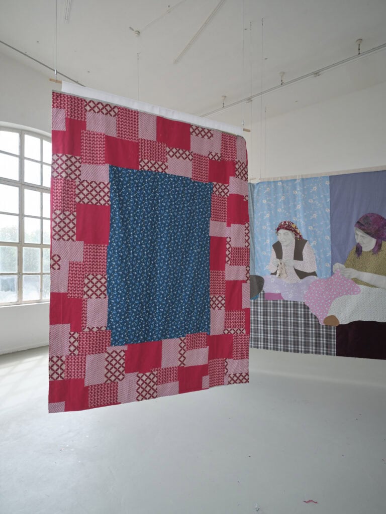 Foto: Aufnahme eines Ausstellungsraums mit weißen Wänden, grauem Boden und einem großen Fenster. Ein großformatiges Textilbild hängt von der Decke abgehängt. Das Bild zeigt zwei weibliche Personen, die auf dem Boden Sitzen und Handarbeiten machen. Davor hängt ein weiteres Textilbild aus geometrischen roten und blauen Flächen.