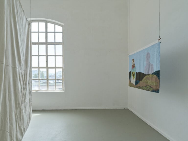Foto: Aufnahme eines Ausstellungsraums mit weißen Wänden, grauem Boden und einem großen Fenster. Vor der rechten Wand hängt ein großformatiges Textilbild. Das Bild zeigt eine weibliche Person. Am linken Rand hängt eine weiße Textilbahn.