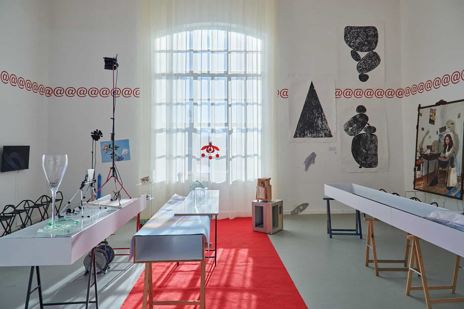 Foto: Ansicht eines Raumes mit weißen Wänden, grauem Boden und großem Fenster. Vor dem Fenster hängen halbtransparente, weiße Vorhänge bis kurz über dem Boden. An der Wand des Raumes befinden sich auf unterschiedlichen Höhen Wandbeklebungen, Papierarbeiten und Bildschirme. Im Raum befinden sich Stühle sowie Skulpturen und Konstruktionen auf Böcken.