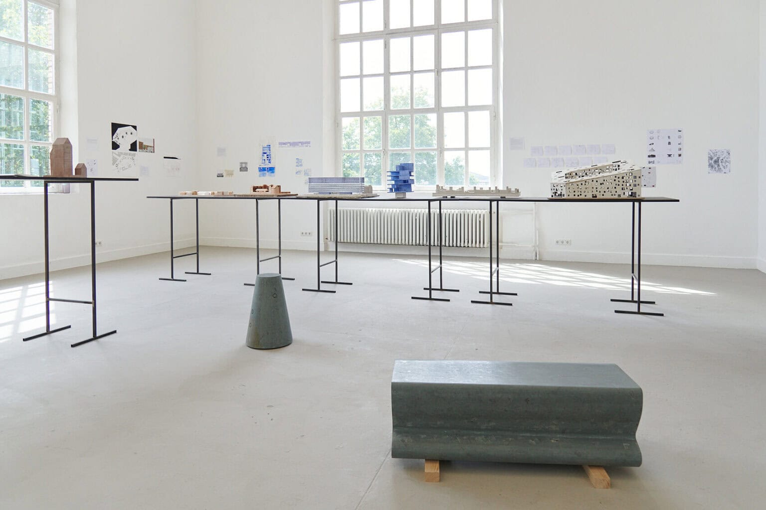Foto: Ein Raum mit weißen Wänden, grauem Boden und zwei Fenstern. An den Wänden befinden sich kleinformatige Papierarbeiten. Im Raum stehen zwei Tische, auf denen sich architektonische Modelle befinden. Auf dem Boden liegen Objekte.