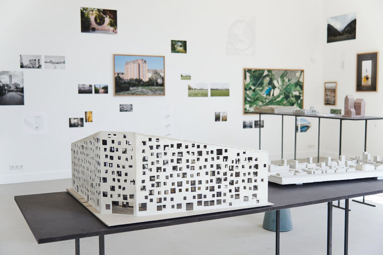 Foto: Ein Raum mit weißen Wänden und grauem Boden. An den Wänden befinden sich gerahmt und ungerahmte, flache Objekte und Klein- und Mittelformaten. Im Raum stehen zwei Tische, auf denen sich architektonische Modelle befinden.