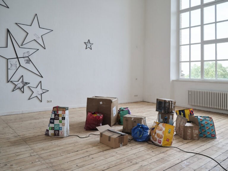 Foto: Aufnahme eines Ausstellungsraums mit weißen Wänden, Holzdielen und einem großen Fenster. In der Mitte des Raums steht eine Installation auf dem Boden bestehend aus mehreren Pappkartons und Tüten. An der linken Wand sind unterschiedlich große schwarze Sterne montiert.