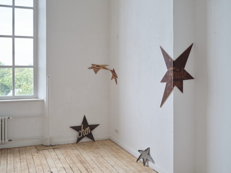 Foto: Aufnahme eines Ausstellungsraums mit weißen Wänden, Holzdielen und einem großen Fenster. An der linken Wand sind unterschiedlich große Sterne aufgehängt oder aufgestellt.