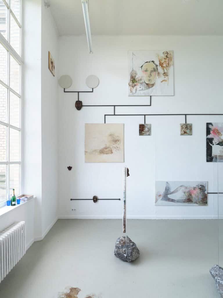 Foto: Aufnahme eines Ausstellungsraums mit weißen Wänden, grauem Boden und einem großen Fenster. Im Raum ist eine mehrteilige Boden- und Wandinstallation aufgebaut, bestehend aus Gemälden, Masken, Steinen und braun fleckigen Farbflächen auf dem Boden.