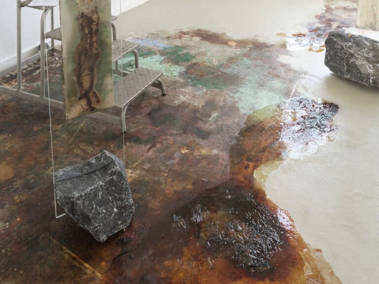 Foto: Detailaufnahme einer Kunstinstallation bestehend aus großen Steinen auf grauem Boden. Unterschiedlich braun fleckige Farbflächen wurden auf dem Boden aufgetragen.