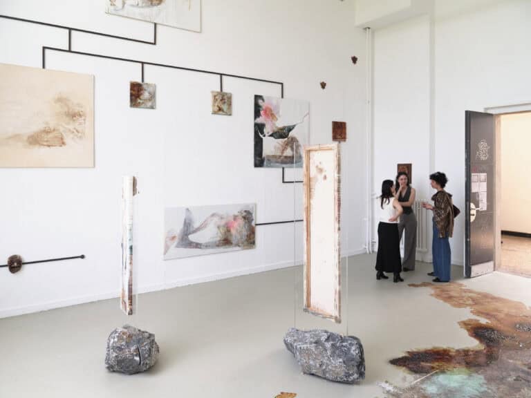 Foto: Aufnahme eines Ausstellungsraums mit weißen Wänden, grauem Boden und einer geöffneten Tür. Im Raum ist eine mehrteilige Boden- und Wandinstallation aufgebaut, bestehend aus Gemälden, Masken, Steinen und braun fleckigen Farbflächen auf dem Boden. Drei Personen stehen an der Tür im Gespräch.