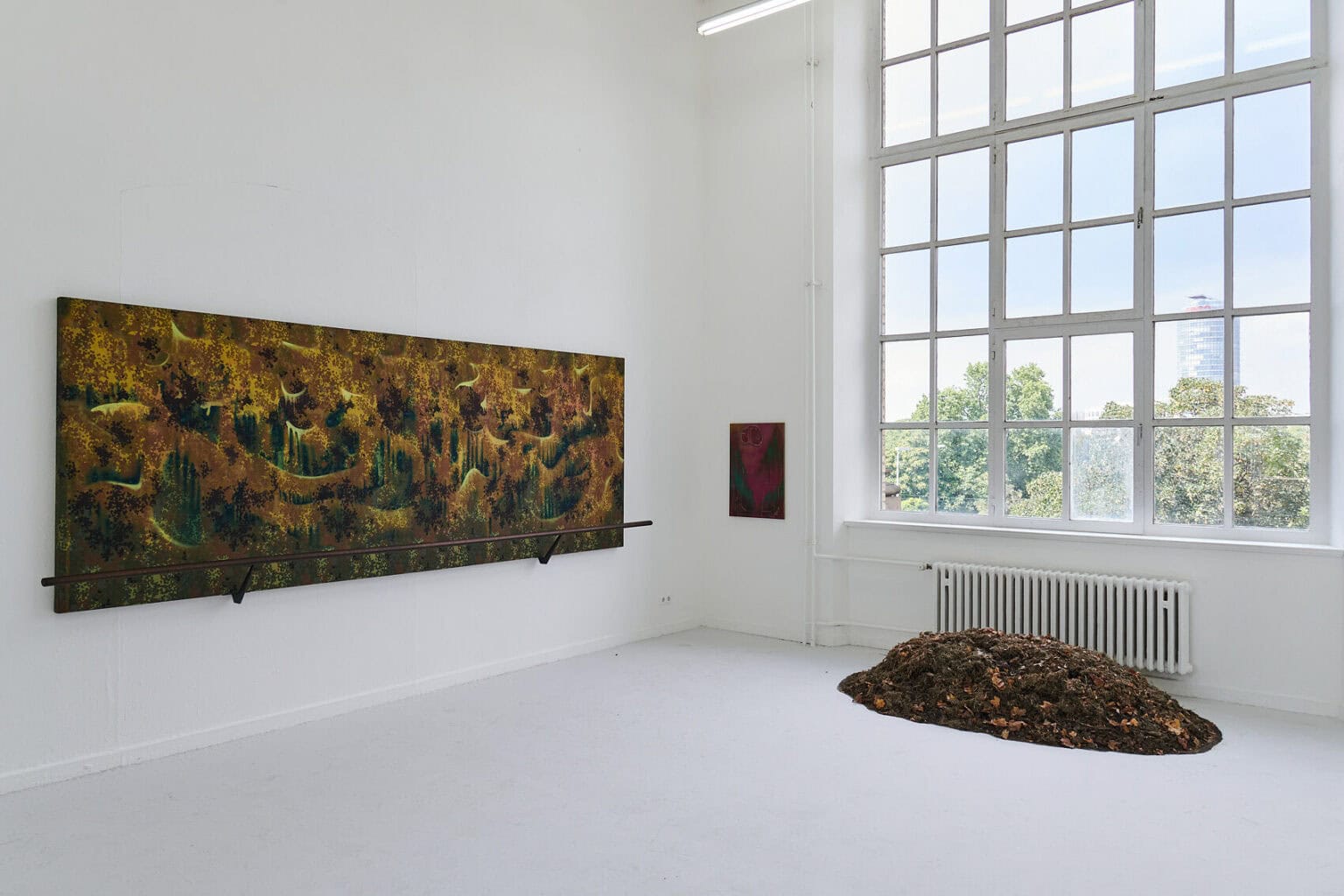 Foto: Raum mit weißen Wänden, grauem Boden und großem Fenster. An den Wänden befinden sich Leinwandarbeiten und Objekte in Klein- und Großformaten. Vor dem Fenster liegt ein Objekt auf dem Boden.