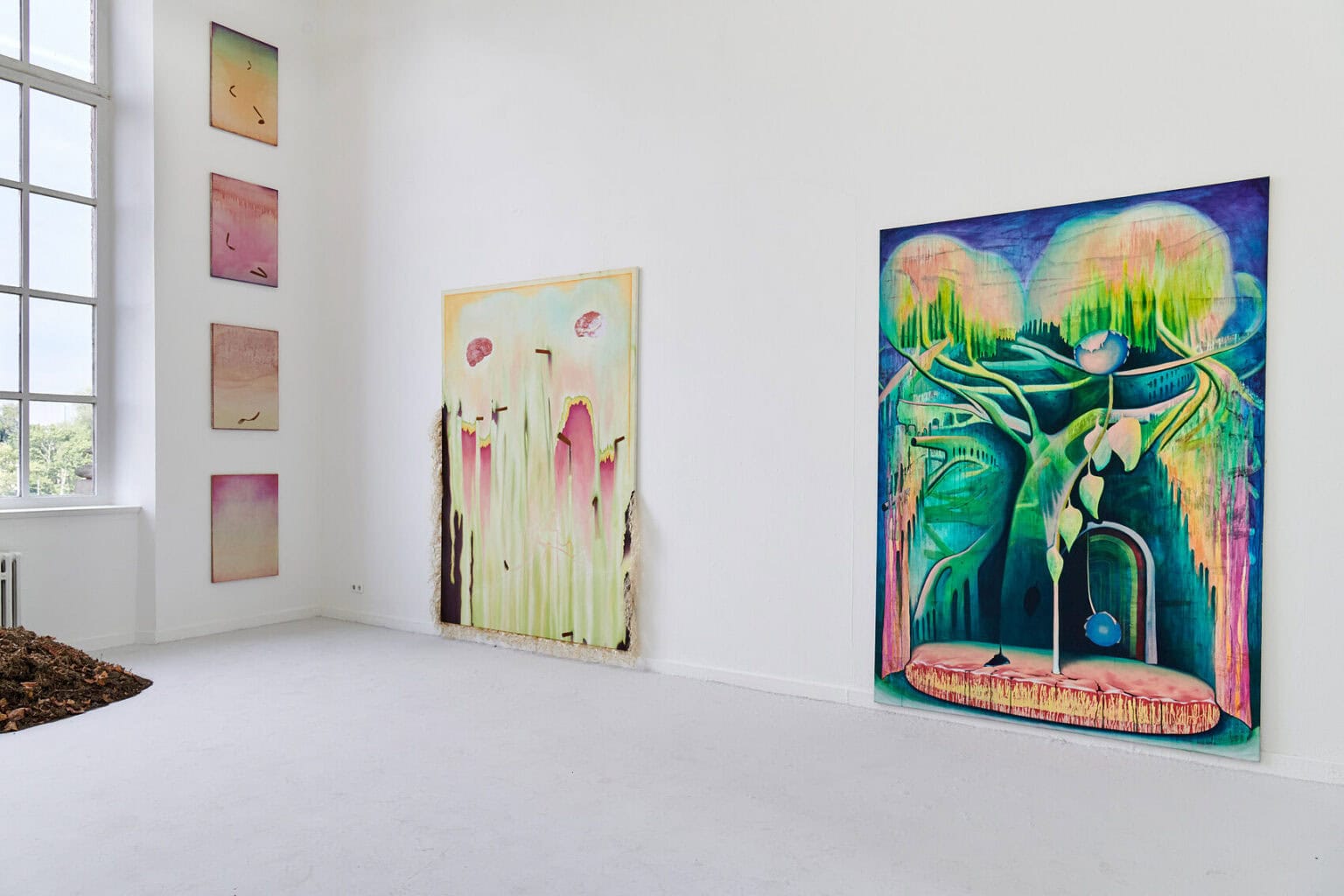 Foto: Raum mit weißen Wänden, grauem Boden und großem Fenster. An den Wänden befinden sich Leinwandarbeiten in Klein- und Großformaten. Vor dem Fenster liegt ein Objekt auf dem Boden.