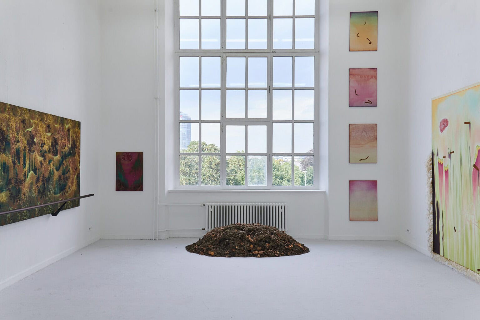 Foto: Raum mit weißen Wänden, grauem Boden und großem Fenster. An den Wänden befinden sich Leinwandarbeiten und Objekte in Klein- und Großformaten. Vor dem Fenster liegt ein Objekt auf dem Boden.