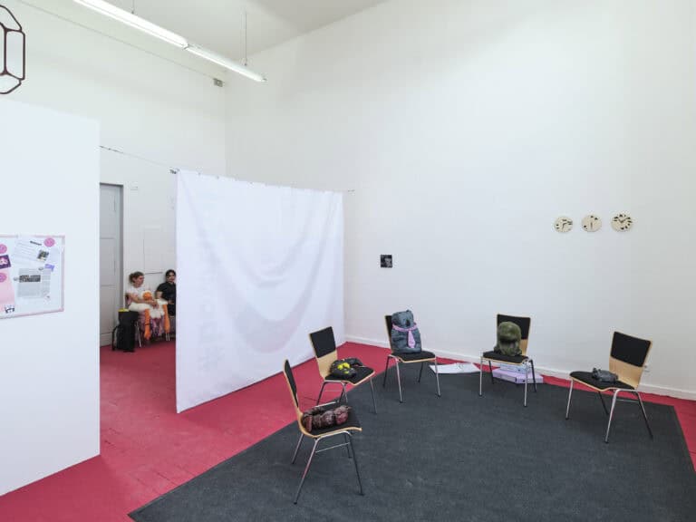 Foto: Aufnahme einer Kunstinstallation in einem hellen Ausstellungsraum mit rotem Boden, auf dem ein dunkelgrauer Teppich liegt. Fünf Stühle sind im Halbkreis aufgestellt und auf jeden Stuhl steht eine Skulptur eines abstrakten Tier- oder Phantasiegeschöpfes. Der linke Raumbereich ist durch ein Textilelement und eine weiße Wand abgetrennt. Zwei Personen sitzen im Hintergrund.