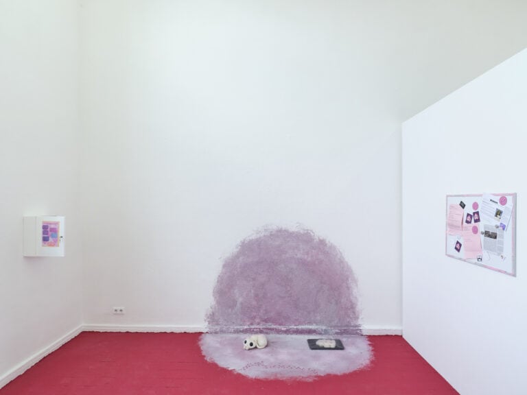 Foto: Aufnahme einer Kunstinstallation in einem hellen Ausstellungsraum mit rotem Boden. Eine hell-violette kreisförmige Farbfläche ist auf Wand und Boden aufgemalt. Unterschiedliche Kunstwerke liegen bzw. hängen an Wand und Boden.