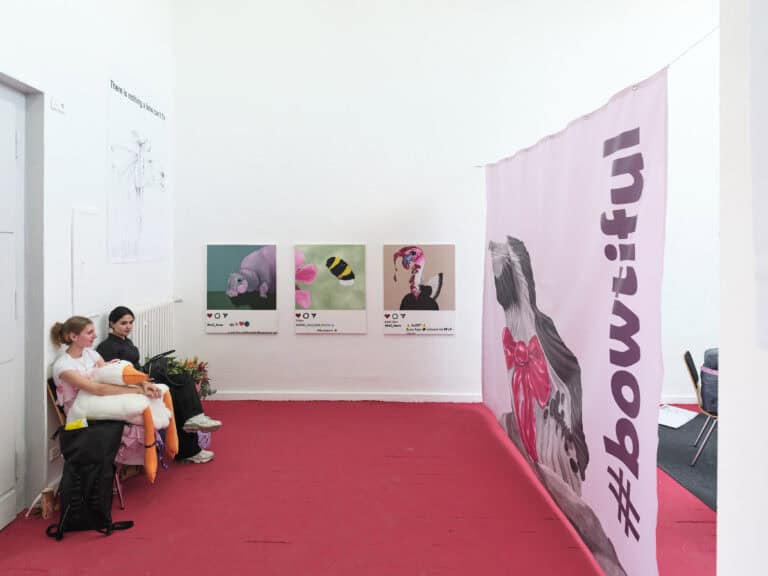 Foto: Aufnahme einer Kunstinstallation in einem hellen Ausstellungsraum mit rotem Boden. Der rechte Raumbereich ist durch ein bedrucktes Banner abgetrennt. An der Rückwand sind drei Drucke aufgehängt.