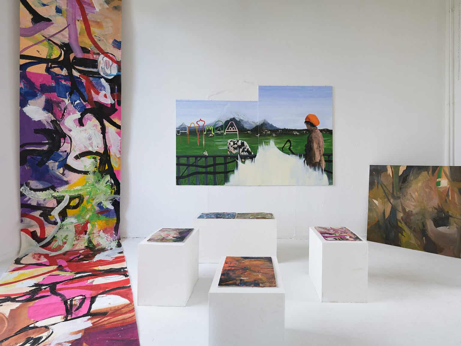 Foto: Ansicht eines Raumes mit weißen Wänden und weißem Boden. Im Raum stehen vier weiße Sockel, auf denen flache Objekte liegen. Eine große, flache, bemalte Rolle hängt in einer der Raumecken von der Decke über den Boden. An der Wand und quer in einer Ecke des hängen zwei Malereien.