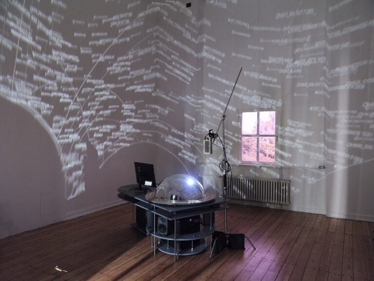Foto: Aufnahme eines abgedunkelten Ausstellungsraums. In der Mitte des Raums steht ein medientechnischer Aufbau. An der Decke zeigen sich Projektionen.