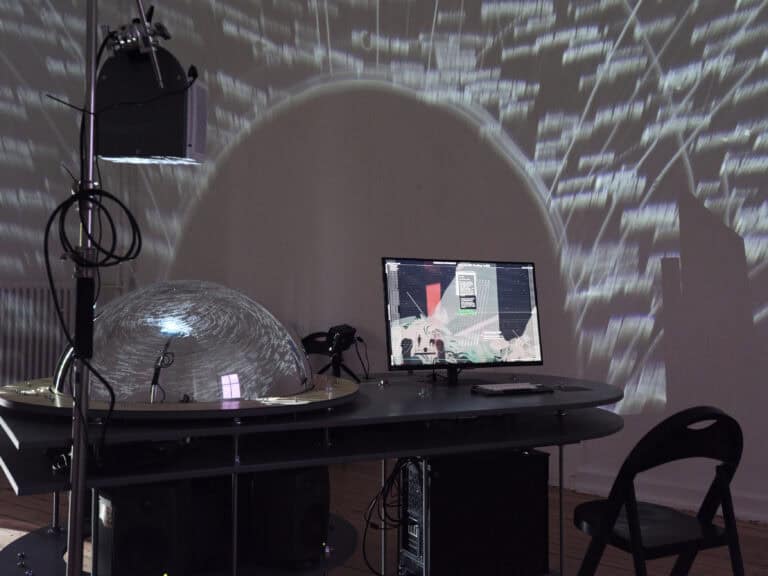 Foto: Aufnahme eines abgedunkelten Ausstellungsraums. In der Mitte des Raums steht ein medientechnischer Aufbau. An der Decke zeigen sich Projektionen.
