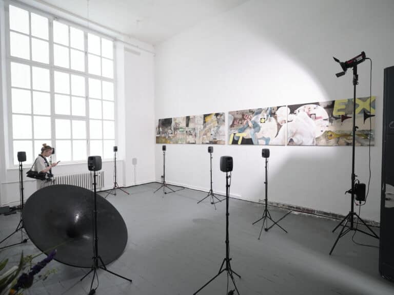 Foto: Aufnahme eines Ausstellungsraums mit weißen Wänden, grauem Boden und einem großen Fenster. Im Raum ist eine mehrteilige Boden- und Wandinstallation aufgebaut. An der Wand ist ein mehrteiliges Gemälde aufgehängt. Auf mehreren Stativen stehen Lautsprecher und Scheinwerfer.