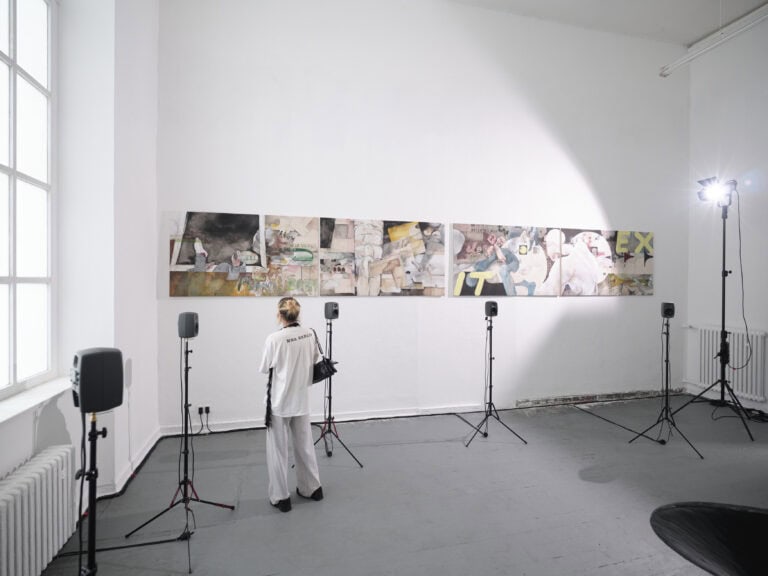 Foto: Aufnahme eines Ausstellungsraums mit weißen Wänden, grauem Boden und einem großen Fenster. Im Raum ist eine mehrteilige Boden- und Wandinstallation aufgebaut. An der Wand ist ein mehrteiliges Gemälde aufgehängt. Auf mehreren Stativen stehen Lautsprecher und Scheinwerfer. Eine Person betrachtet die Arbeiten.