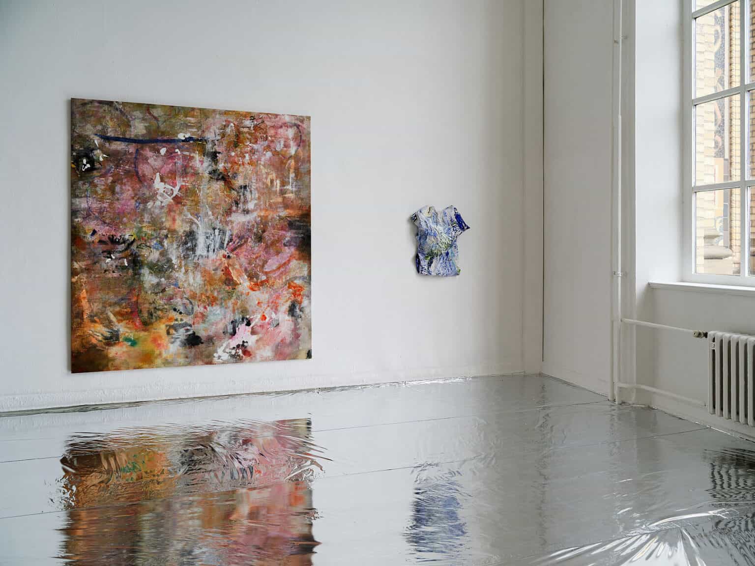 Foto: Ansicht eines Raumes mit weißen Wänden und Fenster. Der Boden ist mit einem silbernen, reflektierendem Material ausgelegt. An der Wand befindet sich ein Objekt und eine großformatige Leinwand. Die Objekte spiegeln sich in der Oberfläche des Bodens.