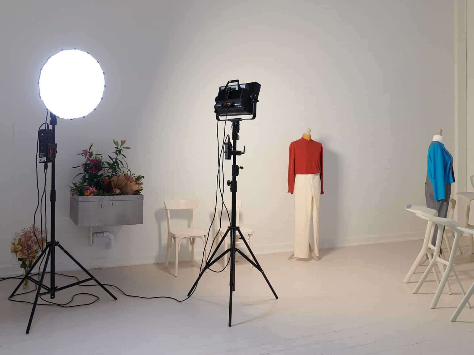 Foto: Ansicht eines Raumes mit weißen Wänden und beigem Boden. An einer der Wände befindet sich ein mit Blumen gefülltes Waschbecken. Im Raum stehen zwei leuchtende Lichter, die auf ausgefahrenen Stativen angebracht sind. Im Raum stehen außerdem mehrere beige Stühle und zwei bekleidete Schneiderpuppen.