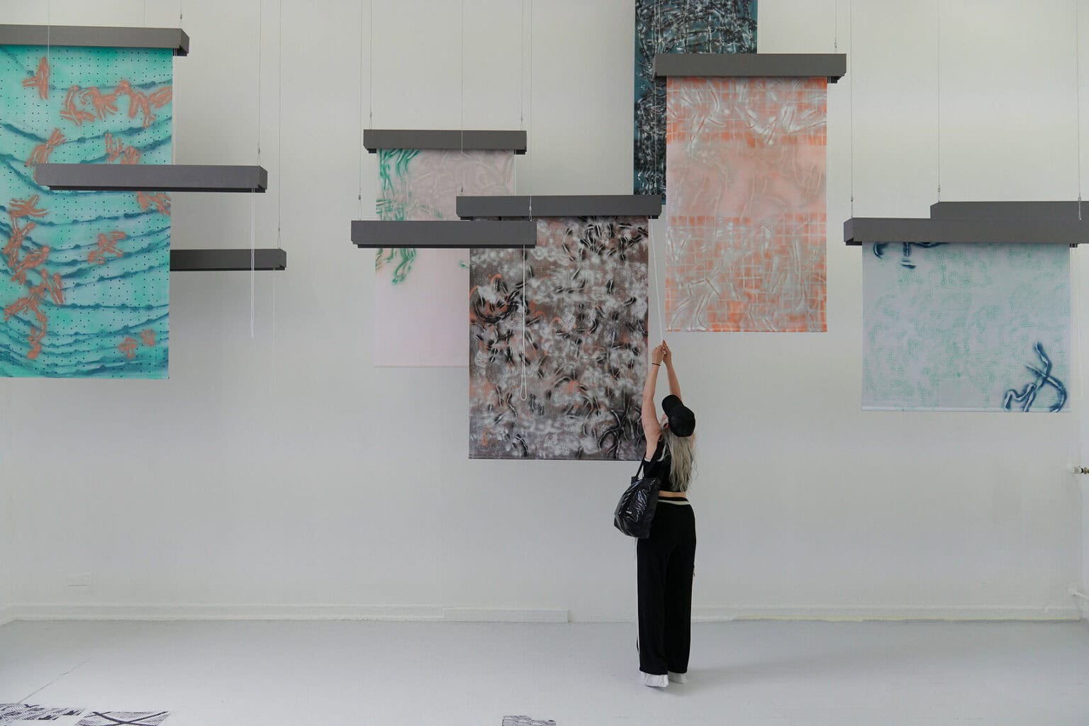 Foto: Ein Raum mit weißen Wänden und grauem Dielenboden. Von der Decke hängen Fäden, an denen flache, herabhängenden Objekte befestigt sind. Auf dem Boden liegt eine Gruppe flacher, gemusterter Objekte. Links im Raum stehen zwei großformatige Leinwandarbeiten, die auf einem mit Rollen versehenen Gerüst stehen. Im Raum steht eine Person, die mit einer der Arbeiten interagiert.
