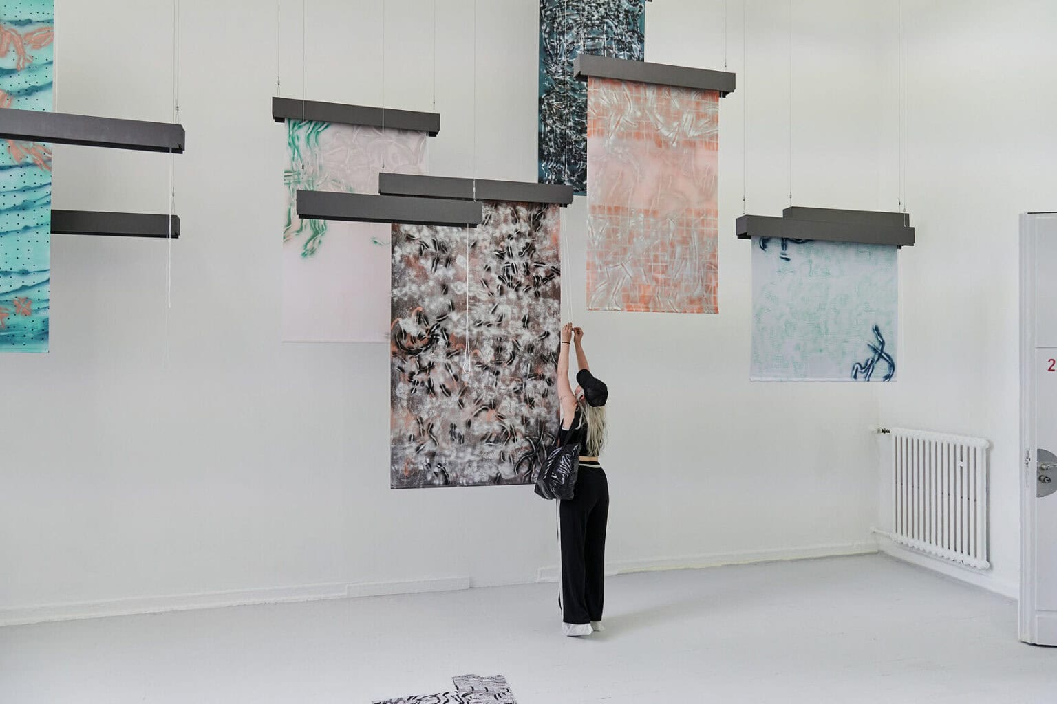 Foto: Ein Raum mit weißen Wänden und grauem Dielenboden. Von der Decke hängen Fäden, an denen flache, herabhängenden Objekte befestigt sind. Auf dem Boden liegt eine Gruppe flacher, gemusterter Objekte. Links im Raum stehen zwei großformatige Leinwandarbeiten, die auf einem mit Rollen versehenen Gerüst stehen. Im Raum steht eine Person, die mit einer der Arbeiten interagiert.