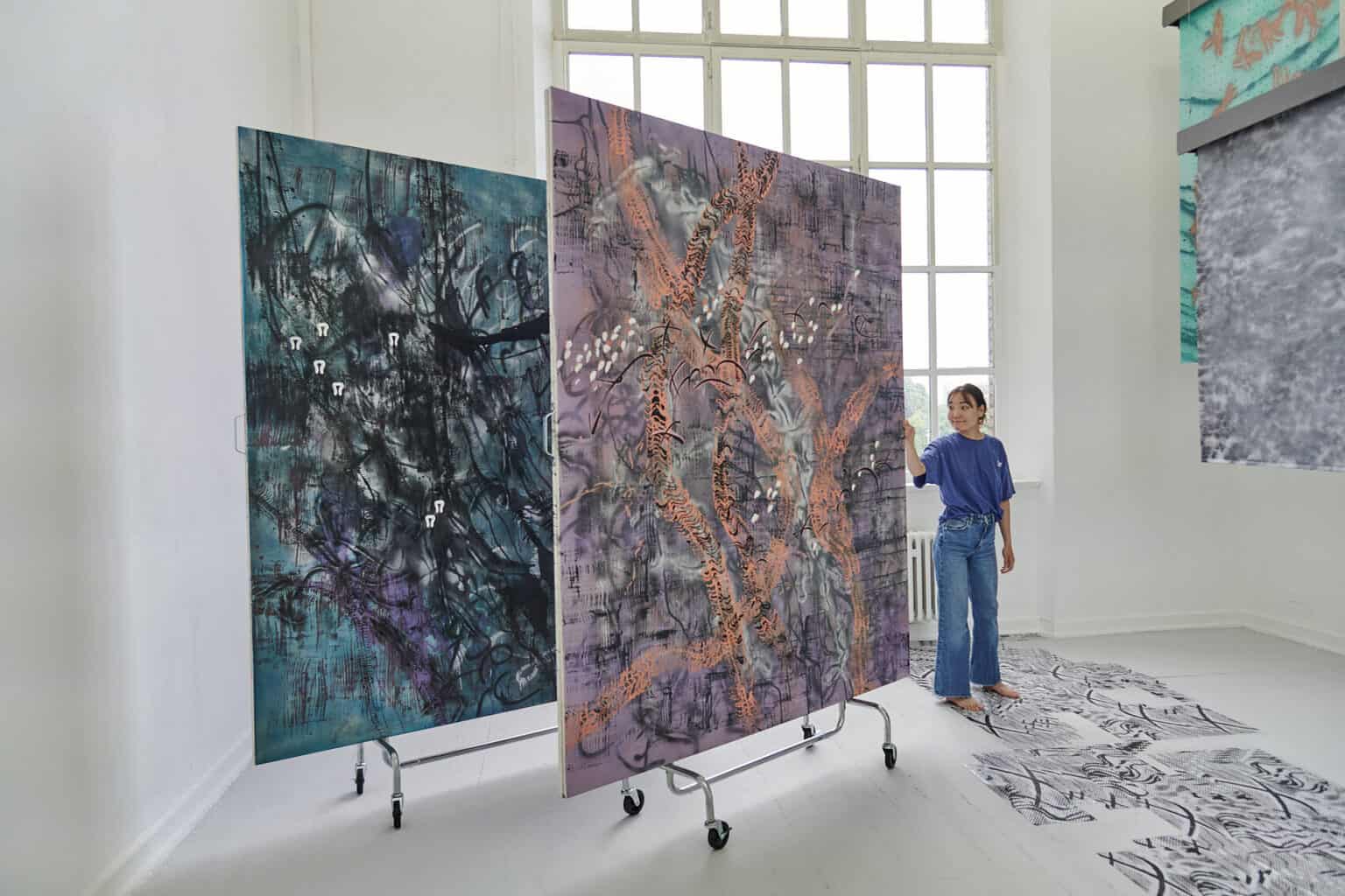 Foto: Ein Raum mit weißen Wänden, grauem Dielenboden und einem großen Fenster. Von der Decke hängen Fäden, an denen flache, herabhängenden Objekte befestigt sind. Auf dem Boden liegt eine Gruppe flacher, gemusterter Objekte. Links im Raum stehen zwei großformatige Leinwandarbeiten, die auf einem mit Rollen versehenen Gerüst stehen. Im Raum steht eine Person.