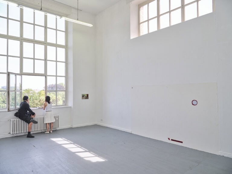 Foto: Aufnahme eines Ausstellungsraums mit weißen Wänden, grauem Boden und großen Fenstern. An den Wänden hängen abstrakte Gemälde. Zwei Personen stehen am offenen Fenster.